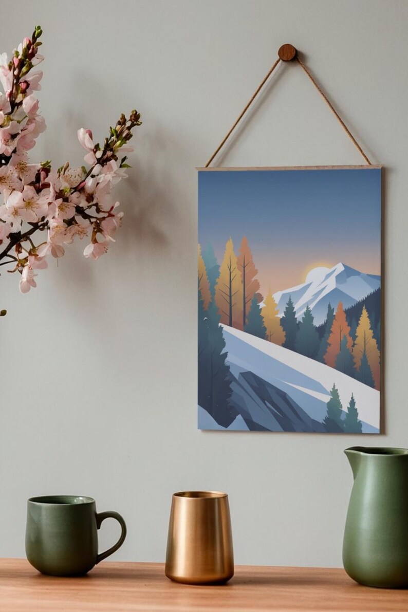 May include: A framed wall art print featuring a mountain landscape with snow-capped peaks, evergreen and autumn-colored trees, and a sunset. The print is suspended by a wooden dowel and a natural cord. A green mug, a gold-colored vase, and a green pitcher are on a wooden surface.