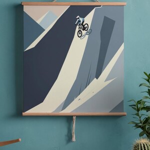 May include: A framed print featuring a mountain biker descending a steep slope. The artwork uses shades of blue, gray, and beige to depict the mountain and rider. The print is mounted on wooden bars and suspended by a rope.