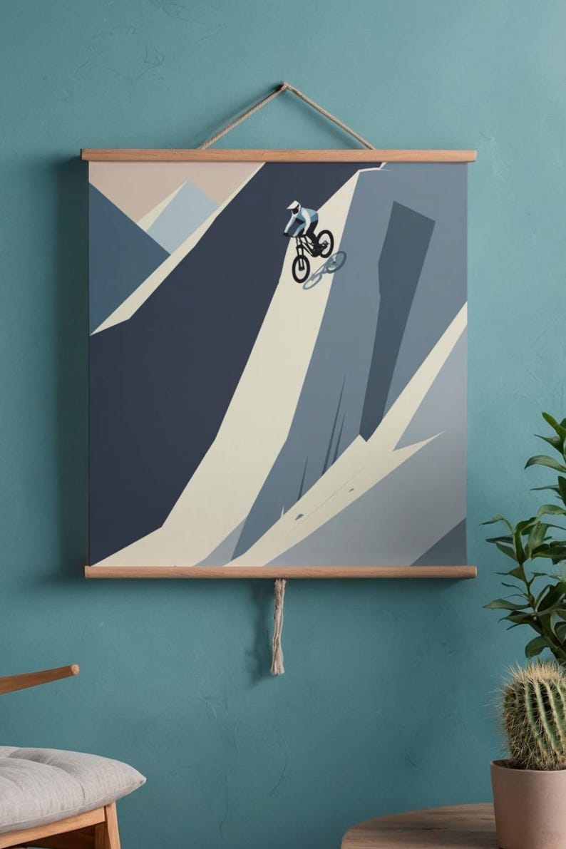 May include: A framed print featuring a mountain biker descending a steep slope. The artwork uses shades of blue, gray, and beige to depict the mountain and rider. The print is mounted on wooden bars and suspended by a rope.