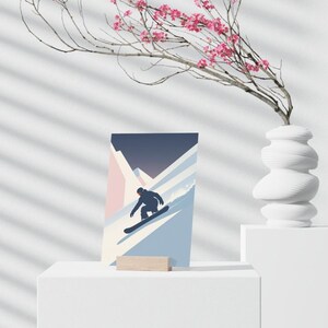 May include: A minimalist art print featuring a snowboarder carving down a mountain slope. The artwork uses a palette of blue, pink, and white, with a dark blue sky. The print is displayed on a white pedestal with a wooden stand.