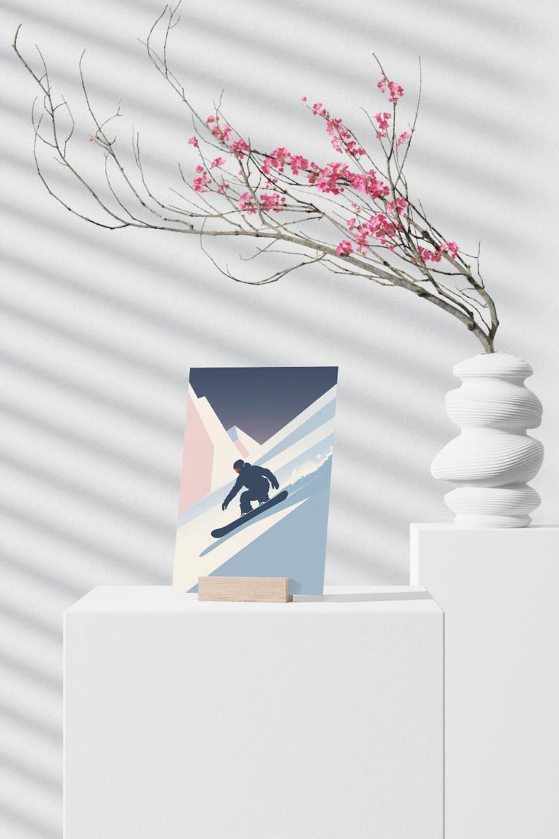 May include: A minimalist art print featuring a snowboarder carving down a mountain slope. The artwork uses a palette of blue, pink, and white, with a dark blue sky. The print is displayed on a white pedestal with a wooden stand.