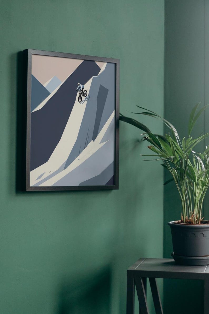 May include: Framed art print featuring a mountain biking scene. The artwork depicts a cyclist navigating a steep, rocky terrain rendered in shades of blue, gray, and beige. The print is encased in a black frame and mounted on a green wall.