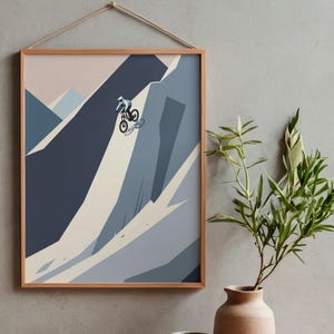 May include: A framed print depicts a mountain biker descending a steep, rocky slope. The artwork uses a minimalist style with shades of blue, gray, and beige. The frame is a light brown color, and the print is hanging on a gray wall.