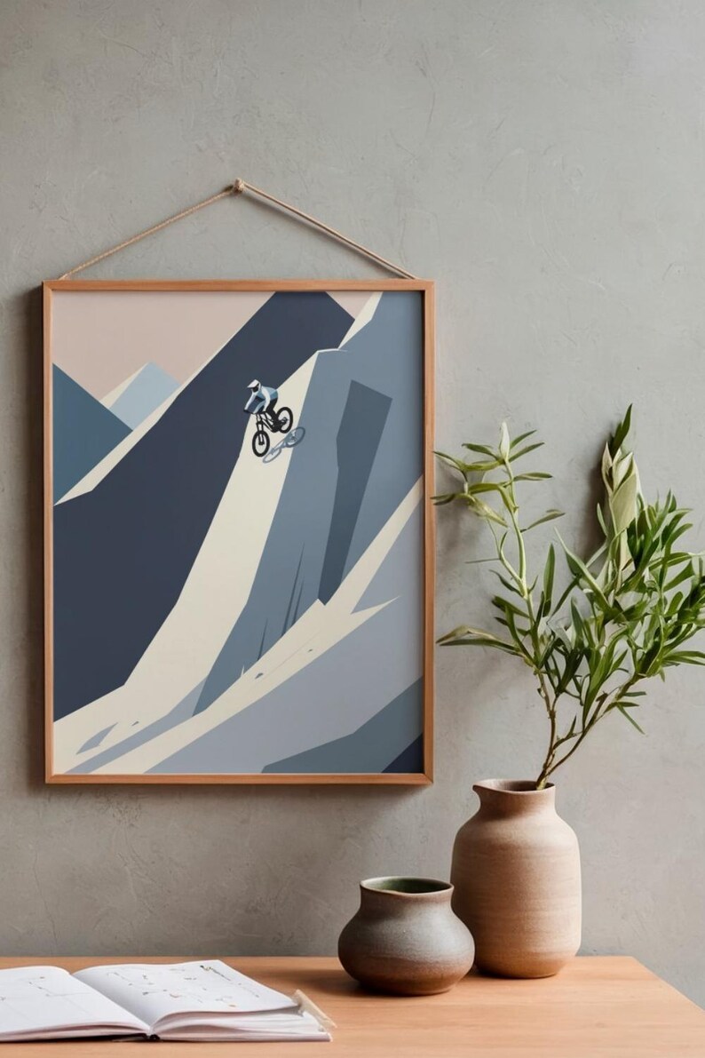 May include: A framed print depicts a mountain biker descending a steep, rocky slope. The artwork uses a minimalist style with shades of blue, gray, and beige. The frame is a light brown color, and the print is hanging on a gray wall.