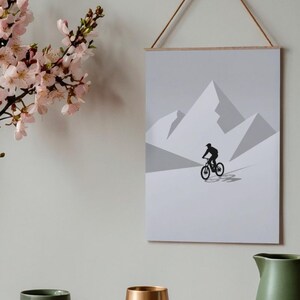 May include: A framed print featuring a minimalist mountain biking scene. The artwork depicts a cyclist on a mountain bike against a backdrop of geometric mountain peaks in shades of gray. The print is suspended by a wooden dowel and a natural cord.