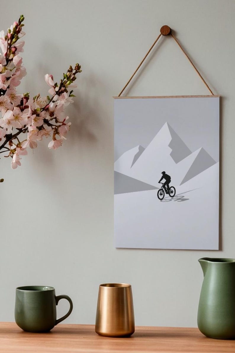 May include: A framed print featuring a minimalist mountain biking scene. The artwork depicts a cyclist on a mountain bike against a backdrop of geometric mountain peaks in shades of gray. The print is suspended by a wooden dowel and a natural cord.