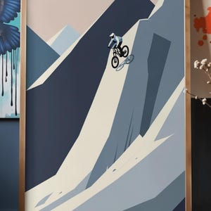 May include: Abstract art print of a mountain biker descending a steep slope. The artwork features a minimalist style with geometric shapes and a muted color palette of blues, beige, and a touch of pink. The framed print is displayed on a wooden surface.