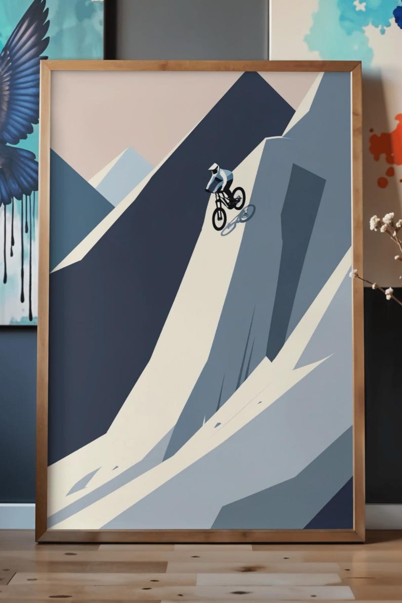 May include: Abstract art print of a mountain biker descending a steep slope. The artwork features a minimalist style with geometric shapes and a muted color palette of blues, beige, and a touch of pink. The framed print is displayed on a wooden surface.