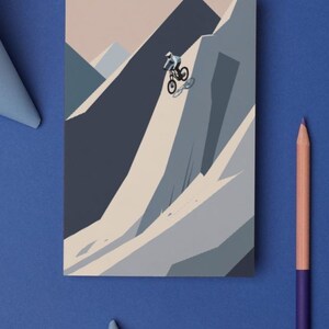 May include: A mountain biking illustration on a rectangular card. The artwork depicts a cyclist descending a steep mountain slope. The color palette includes shades of blue, gray, and beige. A purple and peach colored pencil is on the blue surface.