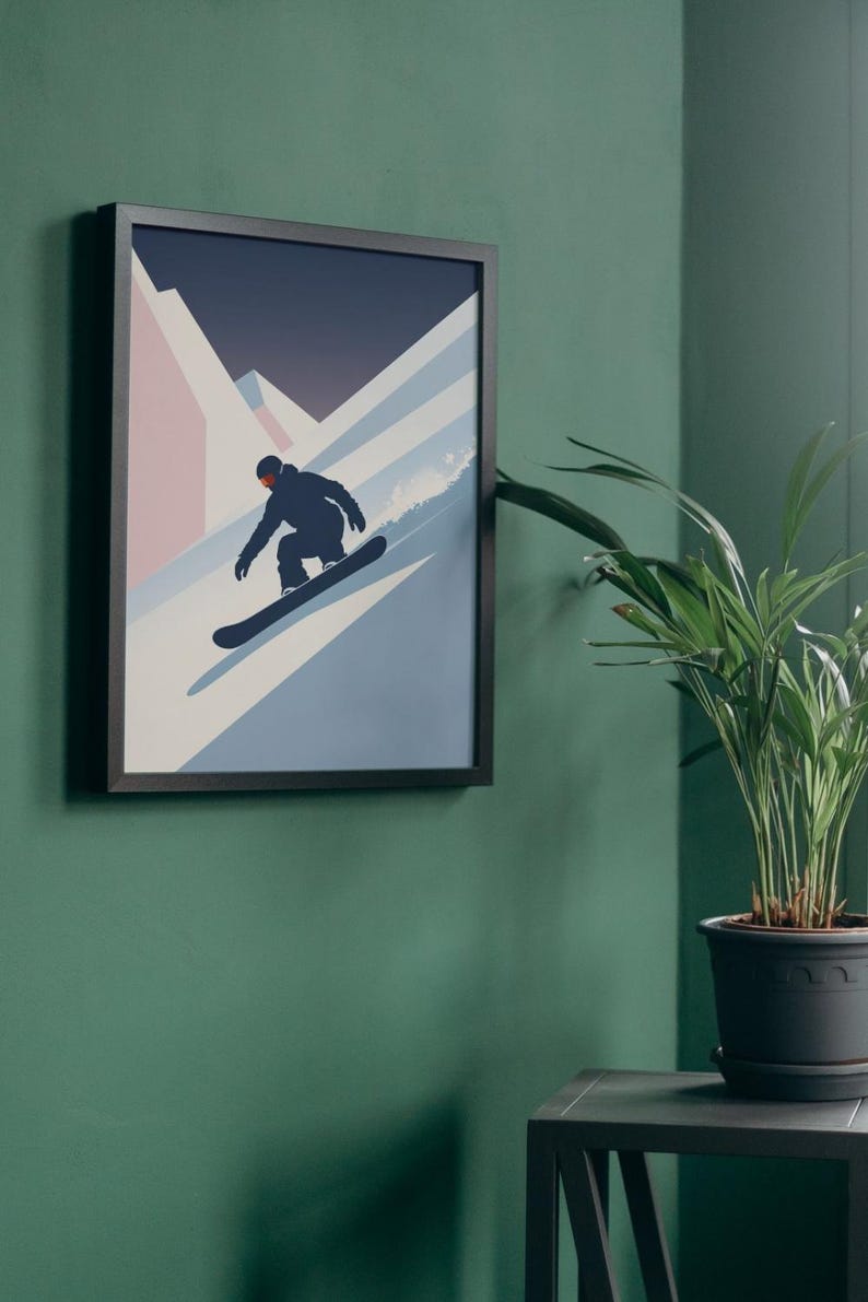 May include: Framed art print featuring a snowboarder in silhouette, descending a snowy slope. The artwork uses a minimalist style with shades of blue, white, and pink against a dark blue sky. The framed print is displayed on a green wall.