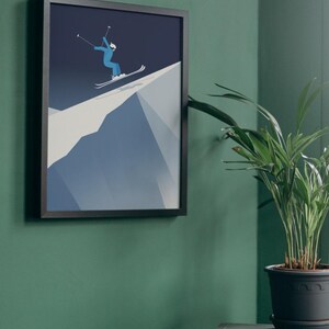 May include: Framed art print featuring a skier in blue attire descending a snowy mountain slope. The artwork is set against a dark blue sky, framed in black, and mounted on a green wall. A potted plant sits nearby.