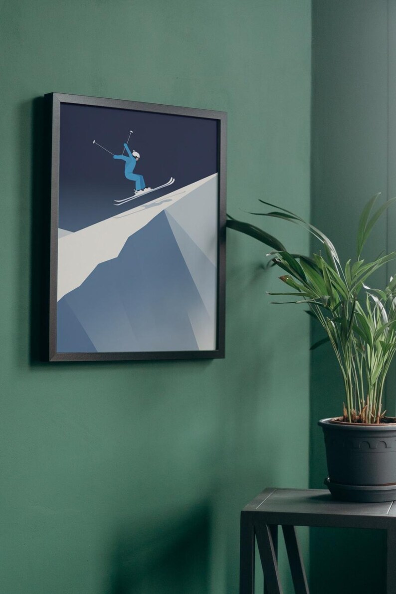 May include: Framed art print featuring a skier in blue attire descending a snowy mountain slope. The artwork is set against a dark blue sky, framed in black, and mounted on a green wall. A potted plant sits nearby.