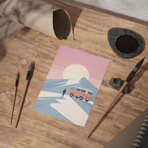 May include: A postcard featuring a retro camper van driving on a snowy mountain road under a large sun. The postcard is on a wooden surface with paint brushes, a notebook, and a stone inkwell.