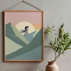 May include: A framed art print featuring a mountain biker silhouetted against a sunset. The artwork uses a minimalist style with shades of green, blue, and peach. The frame is a light wood color, and the print is suspended by a natural rope.