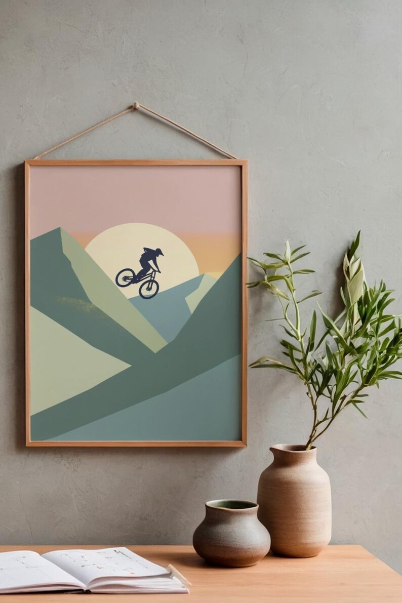 May include: A framed art print featuring a mountain biker silhouetted against a sunset. The artwork uses a minimalist style with shades of green, blue, and peach. The frame is a light wood color, and the print is suspended by a natural rope.
