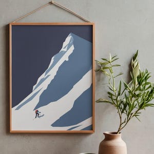 May include: Framed art print featuring a minimalist illustration of a snow-covered mountain with a skier. The artwork is in shades of blue and white, set against a dark blue sky. The print is in a wooden frame and is hung by a natural rope.