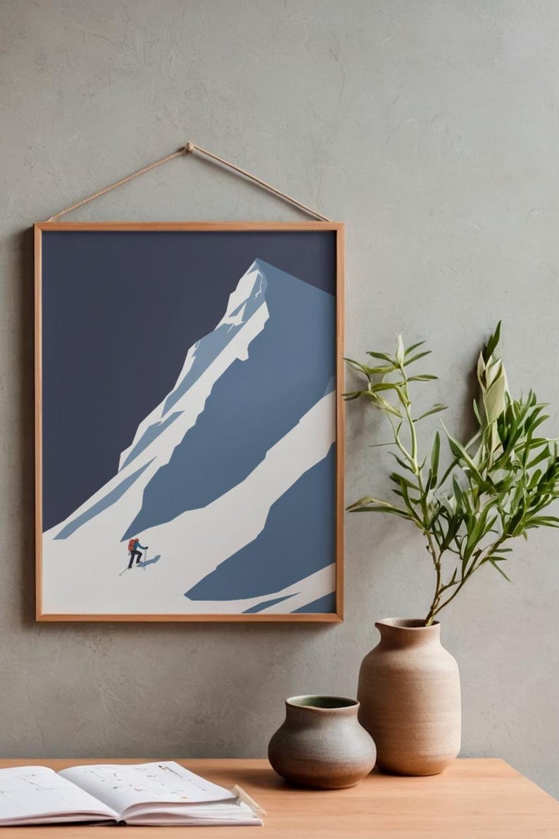 May include: Framed art print featuring a minimalist illustration of a snow-covered mountain with a skier. The artwork is in shades of blue and white, set against a dark blue sky. The print is in a wooden frame and is hung by a natural rope.