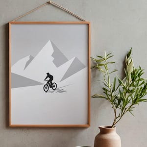 May include: Framed art print featuring a minimalist design of a cyclist riding a mountain bike in front of a mountain range. The artwork is in shades of gray and black, with a natural wood frame and a hanging rope.