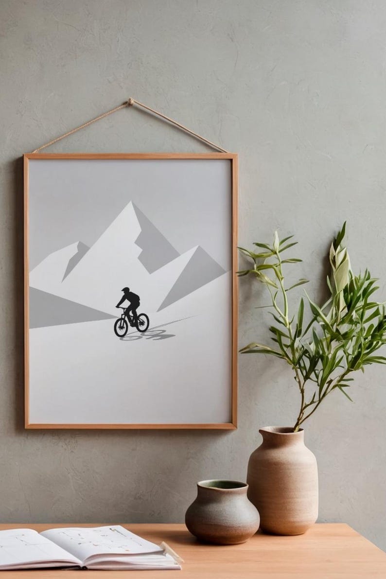 May include: Framed art print featuring a minimalist design of a cyclist riding a mountain bike in front of a mountain range. The artwork is in shades of gray and black, with a natural wood frame and a hanging rope.