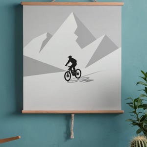 May include: A minimalist poster featuring a black silhouette of a mountain biker descending a slope against a backdrop of gray mountains. The artwork is mounted on wooden bars and suspended by a natural rope.