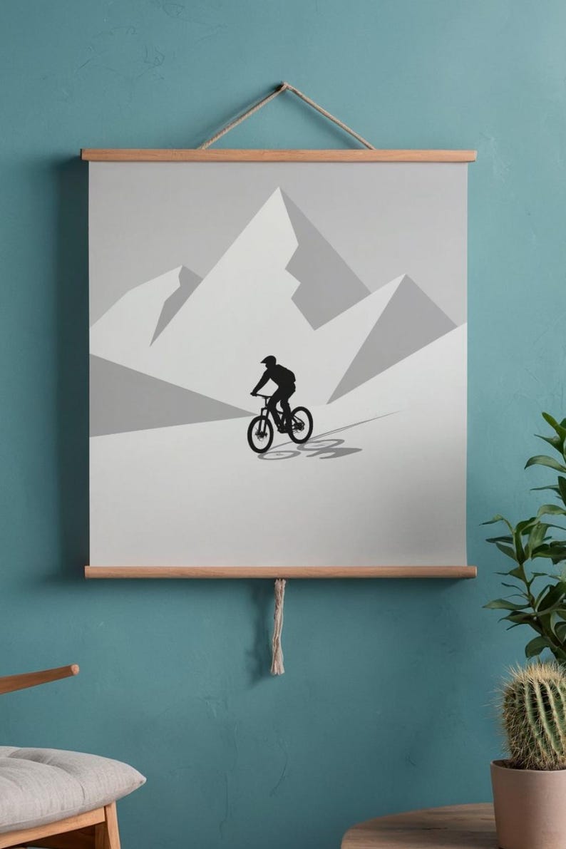 May include: A minimalist poster featuring a black silhouette of a mountain biker descending a slope against a backdrop of gray mountains. The artwork is mounted on wooden bars and suspended by a natural rope.