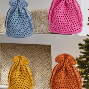 Puede incluir: Cuatro bolsas de cordón de ganchillo en tonos azul, rosa, amarillo y naranja. Las bolsas tienen un diseño de tejido abierto y se recogen en la parte superior con un cierre de cordón. Se exhiben en un estante blanco.