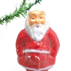 May include: Vintage Santa Claus figurine in a classic red suit with white trim and beard. The figurine has a weathered appearance, with visible paint chipping. A green garland is visible in the upper left corner. A festive Christmas decoration.