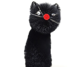 May include: A black cat figurine crafted from fuzzy pipe cleaners, featuring a round head and cylindrical body. It has two small white eyes with black pupils, a bright red circular nose, and thin silver wire whiskers, all mounted on a red base.