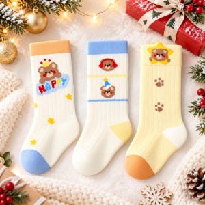 Könnte beinhalten: Drei Paar gerippte Socken mit Bärenmotiven. Die erste ist cremefarben mit orangefarbenem Bündchen, einem Bären mit Krone und dem Wort HAPPY. Die zweite ist cremefarben mit blauem Bündchen, Bärengesichtern und blauen Streifen. Die dritte ist gelb mit Pfotenabdrücken.
