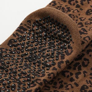 May include: A brown sock featuring a dark brown leopard print pattern. The sole of the sock is covered in small, circular black silicone grips designed to provide traction. The fabric appears to be a soft knit material with a visible texture.