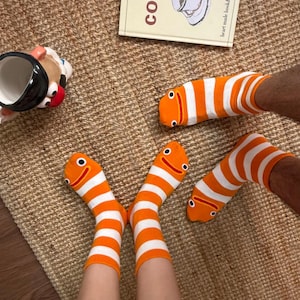 Op de afbeelding: Oranje en wit gestreepte sokken met een cartoon visgezichtsontwerp. De sokken liggen op een geweven bruin tapijt, met een koffieboek en een koffiemok op de achtergrond.