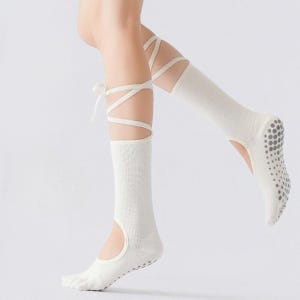 Könnte beinhalten: Weiße Socken im Ballet-Stil mit Riemendesign. Die Socken haben einen Ausschnitt für die Zehen und einen rutschfesten Griff an der Sohle. Die Riemen wickeln sich um Knöchel und Wade und sind mit einer Schleife gebunden.
