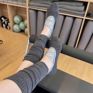 May include: Gray ballet-style socks with light blue criss-cross straps. The socks are paired with gray ribbed leg warmers. The image is taken indoors, with a wooden shelf and exercise equipment in the background.