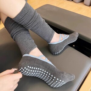 May include: Gray yoga socks with light blue accents and white grip dots on the soles. The socks have a ballet flat design and are paired with matching gray leg warmers. The socks are designed for comfort and grip during exercise.
