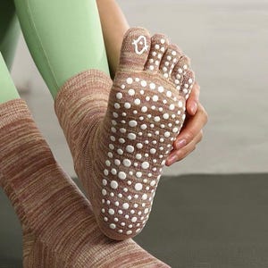 Puede incluir: Calcetines de dedos marrones y beige con puntos de agarre blancos en las suelas. Los calcetines tienen una textura acanalada y un pequeño diseño blanco en el dedo gordo. El texto "SnugCloudDesign" es visible.