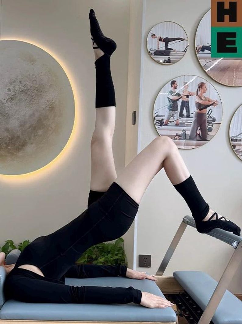 May include: A person wearing black activewear, including a long-sleeved top, shorts, and socks, performing an exercise on a Pilates machine. The background features circular mirrors with exercise demonstrations and a large, illuminated moon-shaped light fixture.