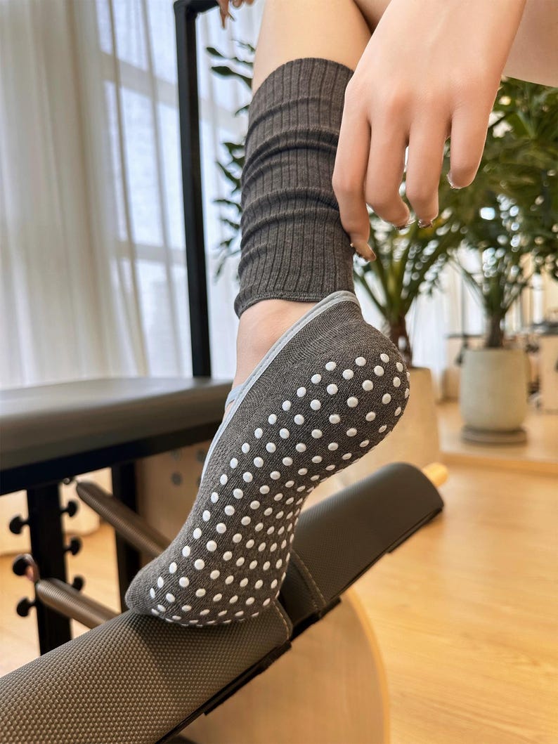 May include: Gray non-slip socks with white dot grips are shown. The socks are worn with gray leg warmers. The socks are designed for activities like yoga or Pilates. The image shows the socks on a piece of exercise equipment.