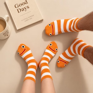May include: Three crew socks featuring horizontal orange and white stripes. Each sock has a cartoon face at the toe with two black eyes and a red mouth. The socks are worn on feet, positioned near a book titled Good Days and a white ceramic mug.