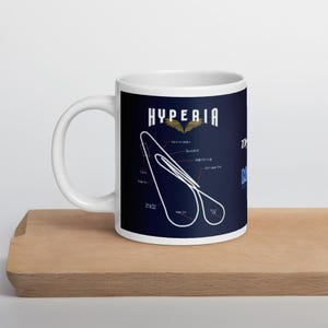Puede incluir: Taza de cerámica blanca con fondo azul oscuro con la palabra "HYPERIA" en blanco, un diseño de pista y texto. La taza está sobre una superficie de madera.