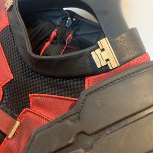 May include: Close-up of a red and black costume with gold accents. The costume features a black collar with a gold clasp, red panels, and a black mesh-like material. The design includes a zipper and a padded shoulder piece.