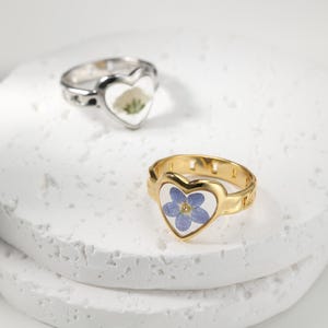 Op de afbeelding: Twee hartvormige ringen. Eén is zilverkleurig met een wit en groen bloemmotief, de andere is goudkleurig met een blauwe bloem. Beide ringen hebben een helder harsmidden, dat de delicate bloemendetails laat zien.