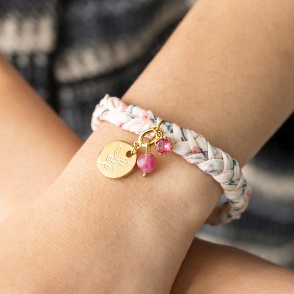 May include: A white fabric bracelet with a floral pattern, braided and tied with a gold charm featuring a leaf design. The charm is attached to a pink gemstone and a smaller pink rhinestone.