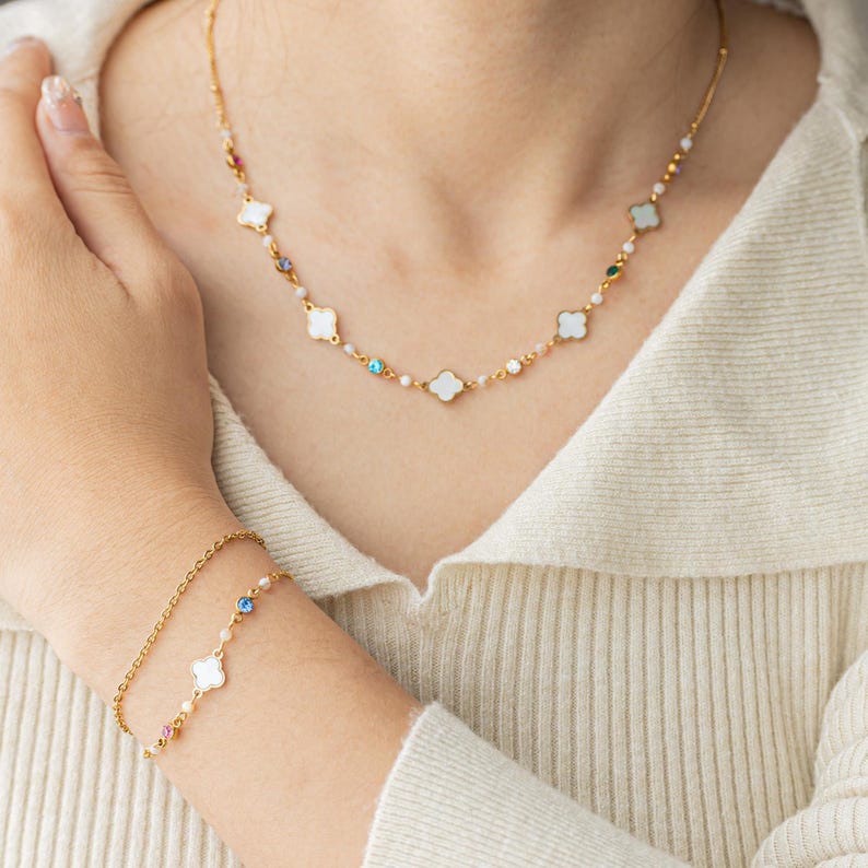 May include: A gold necklace and bracelet set with white clover charms and colorful crystal accents. The necklace features a delicate chain with small white beads and the bracelet has a simple chain with a single clover charm.