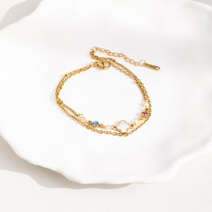May include: A gold-toned layered chain bracelet with a white clover charm and small blue and pink gemstones.