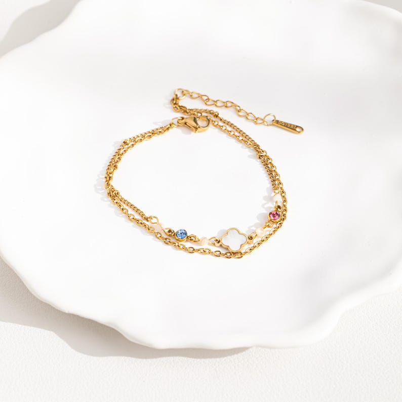 May include: A gold-toned layered chain bracelet with a white clover charm and small blue and pink gemstones.