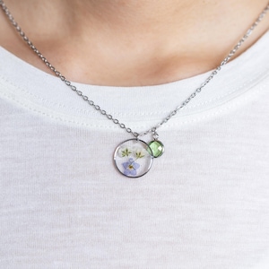 May include: A silver chain necklace with a round pendant featuring pressed flowers and a small green gemstone.