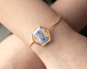 Puede incluir: Pulsera dorada con un colgante hexagonal que contiene flores secas azules y blancas encapsuladas en resina transparente. La delicada cadena conecta con el colgante, creando un accesorio elegante. Una joya única.
