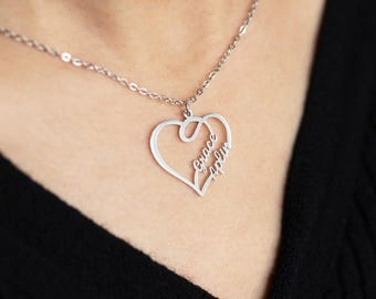 Op de afbeelding: Een zilverkleurige ketting met een hartvormige hanger. De hanger heeft de namen "Grace" en "Aplin" in een sierlijk handschrift. De ketting wordt gedragen tegen een zwarte top, wat het ontwerp van de hanger benadrukt.