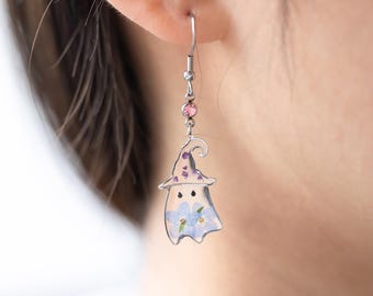 May include: Silver earrings with a clear resin ghost charm featuring pressed flowers. The ghost is wearing a purple and white hat. The earrings have a pink crystal accent.