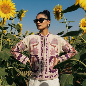 May include: A cream jacket featuring detailed purple and brown floral embroidery. The jacket has a stand-up collar and is worn by a person in a sunflower field. The word "June" is written in cursive at the bottom.